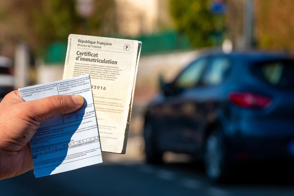Main tenant un certificat d'immatriculation fran&ccedil;ais et une carte internationale d'assurance automobile devant un v&eacute;hicule bleu gar&eacute;