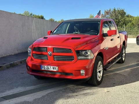 Scène extérieure avec pickup rouge RAM garé sur parking béton, mur gris en arrière-plan, végétation et ciel bleu