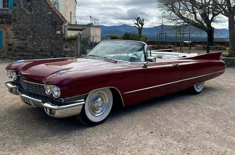 Scène extérieure avec voiture décapotable classique rouge bordeaux stationnée sur terrain gravillonné, bâtiments en pierre et montagnes en arrière-plan, ciel nuageux