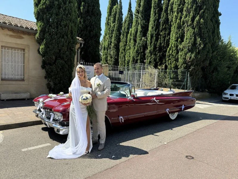 Femme joyeuse sortant d'une voiture rouge décapotable, bras levés en signe de liberté, portant chapeau de paille et chemise bleue, sur route avec paysage montagneux au coucher de soleil